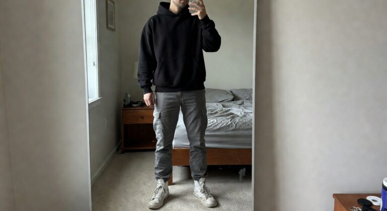 Mirror selfie of a man in an oversized black hoodie, grey cargo pants with multiple pockets, and white high-top sneakers, standing casually in a bedroom