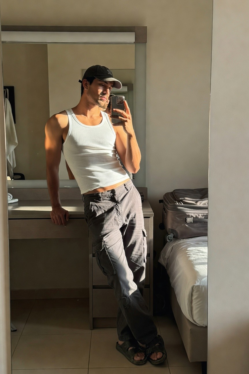 Muscular man in white sleeveless tank top, low-slung grey cargo pants, black cap, and flip-flops, leaning on a dresser in a hotel room mirror selfie with suitcase nearby