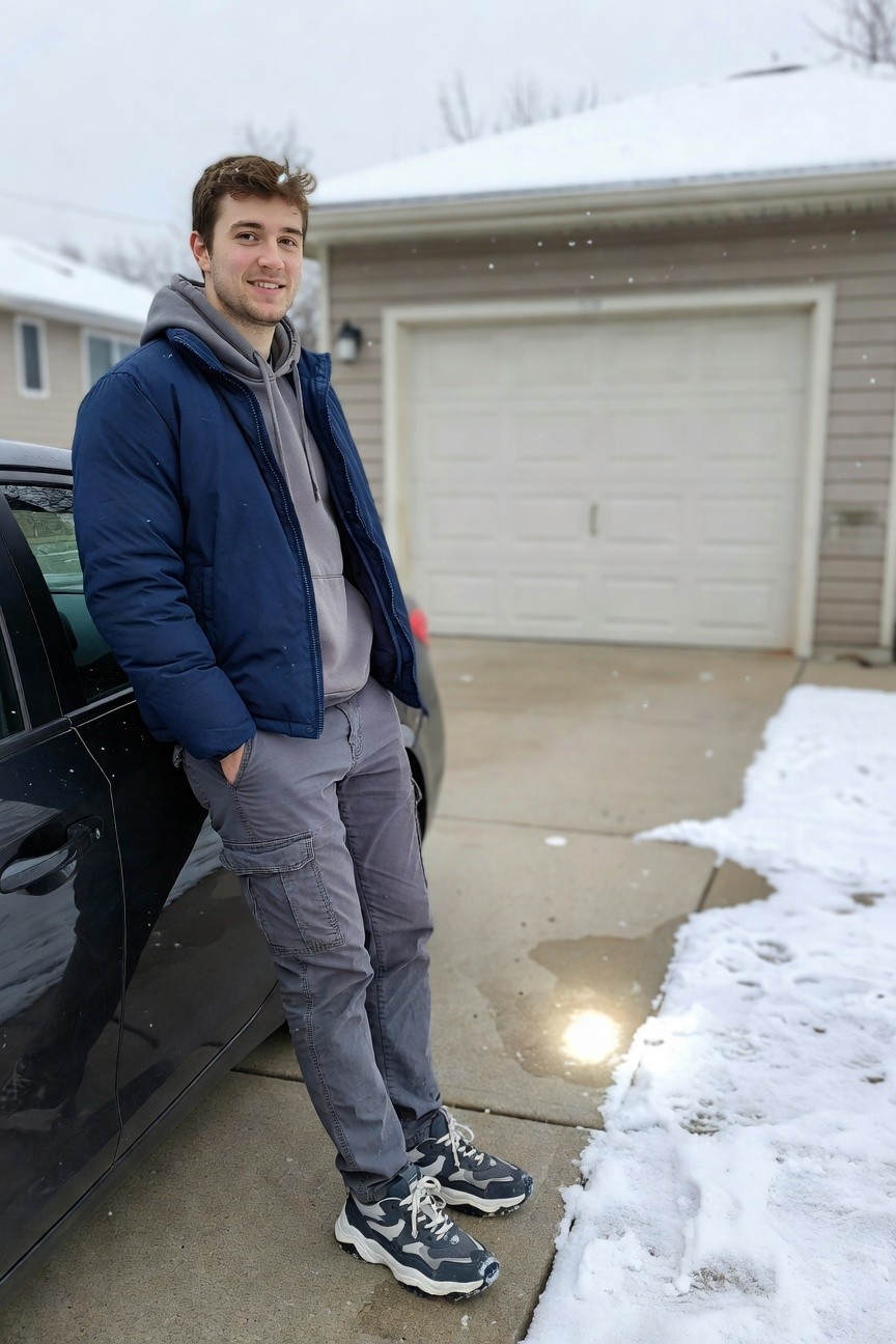 Young man in grey cargo pants, grey hoodie under navy puffer jacket, white and grey sneakers, leaning against black car on snowy driveway