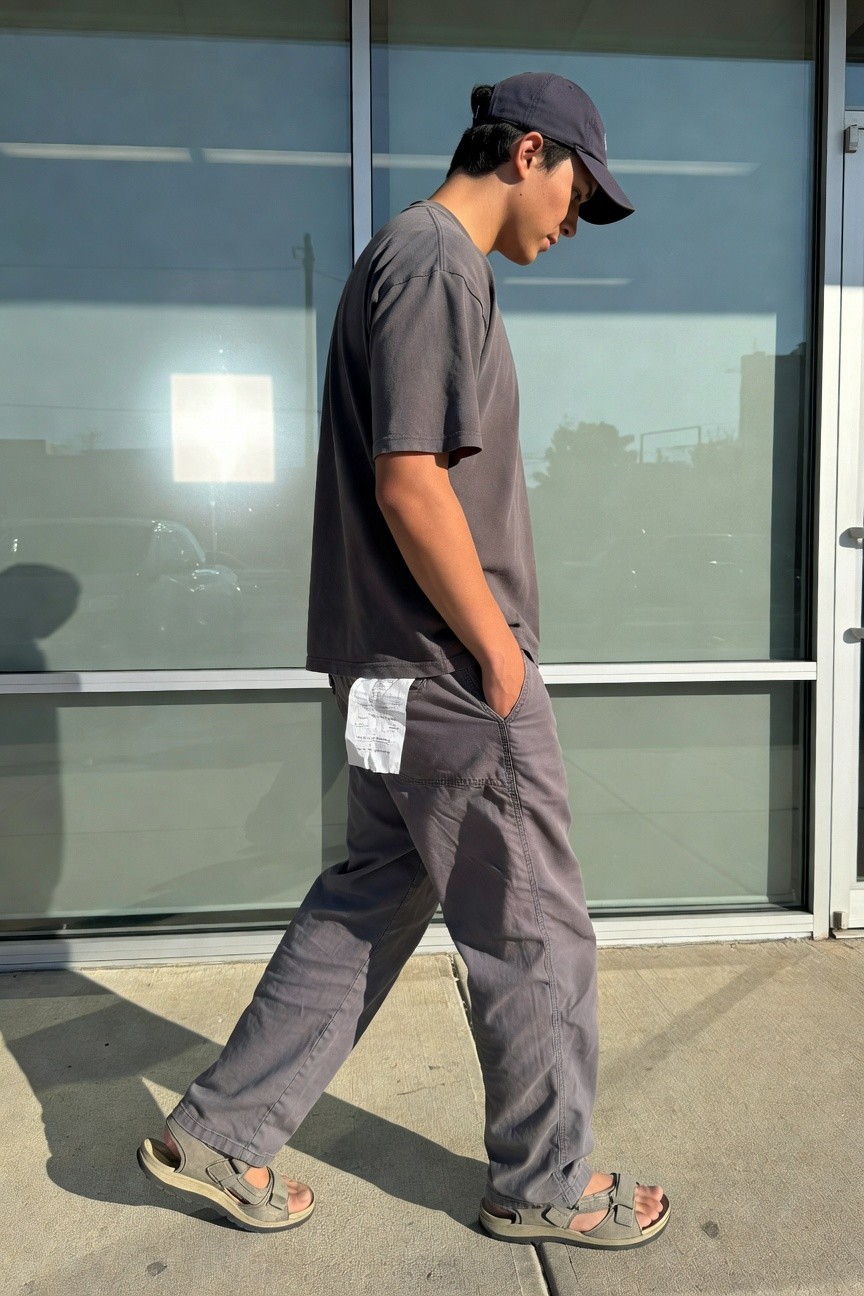 Side view of young Asian man in grey oversized t-shirt, baggy grey cargo pants with dangling receipt from waistband, navy cap, and white strap sandals, walking on concrete sidewalk near glass building