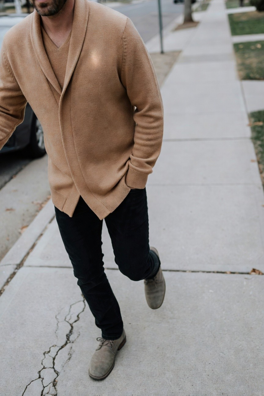 Man walking on sidewalk in open camel knit cardigan over V-neck, black slim jeans, and grey suede derby shoes, hands in pockets for casual cozy style