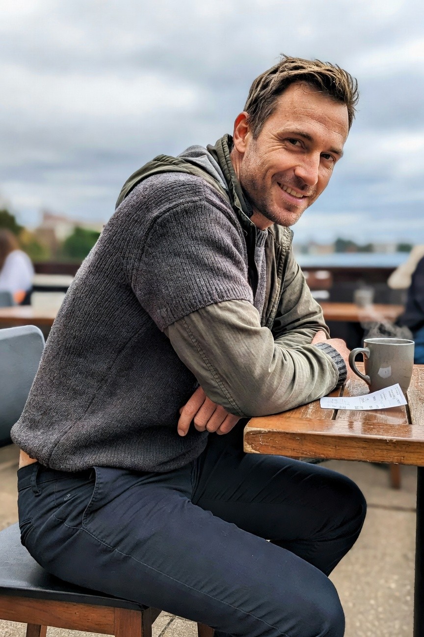Man in gray hooded knit sweater with olive green sleeves and arms, dark navy slim pants, sitting casually at wooden table holding coffee mug