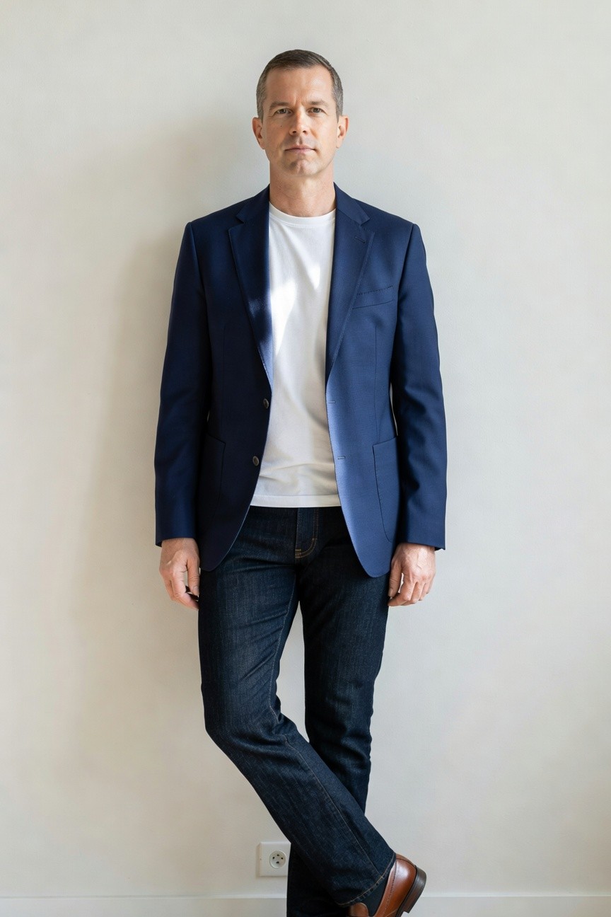 Man standing casually against white wall in navy blazer over white crewneck t-shirt, dark blue jeans, and brown leather dress shoes