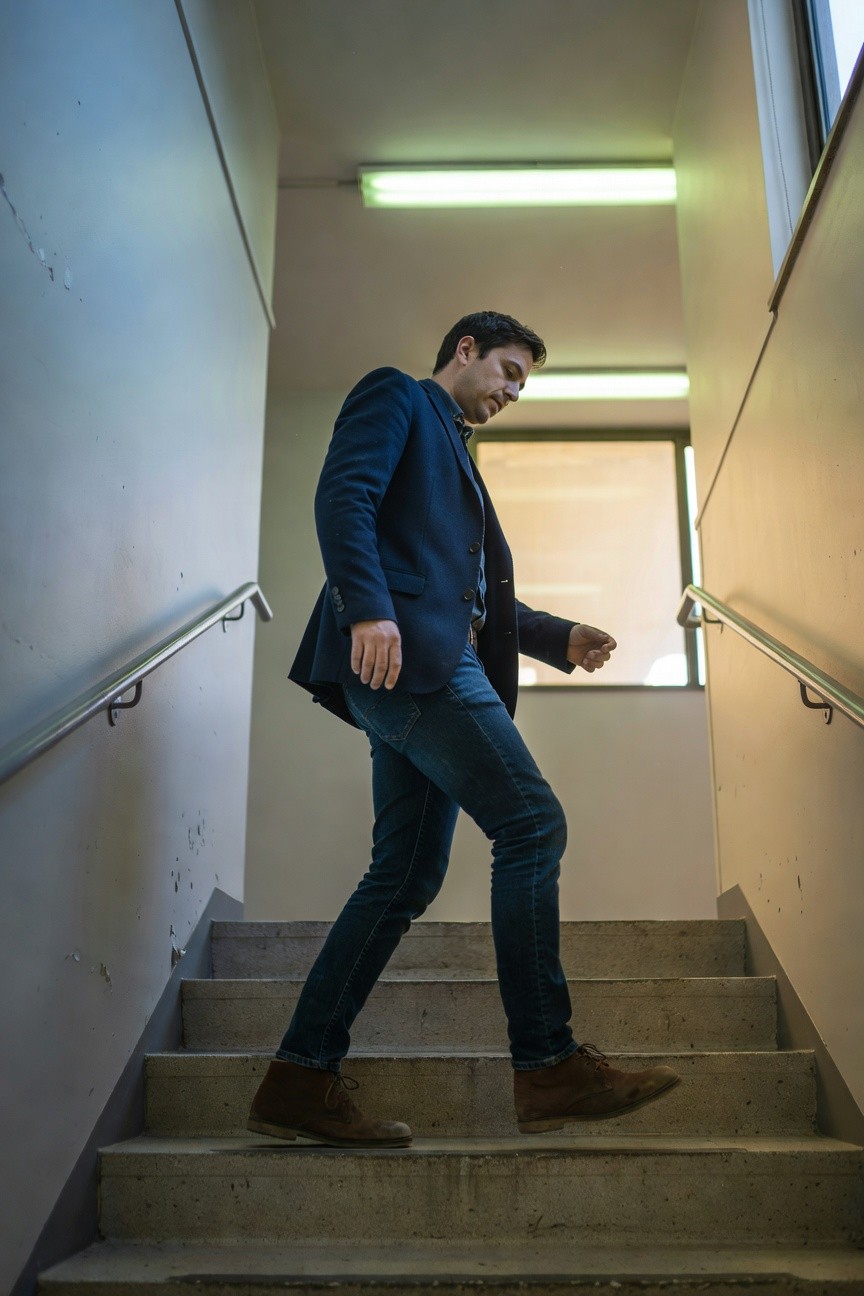 Man walking up stairs in navy blazer over collared shirt, dark slim jeans, and brown suede chukka boots, indoor setting