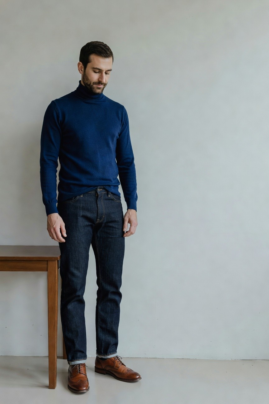 Man in navy turtleneck sweater, dark straight-leg jeans, and brown wingtip brogue shoes standing casually by a wooden table against a plain wall