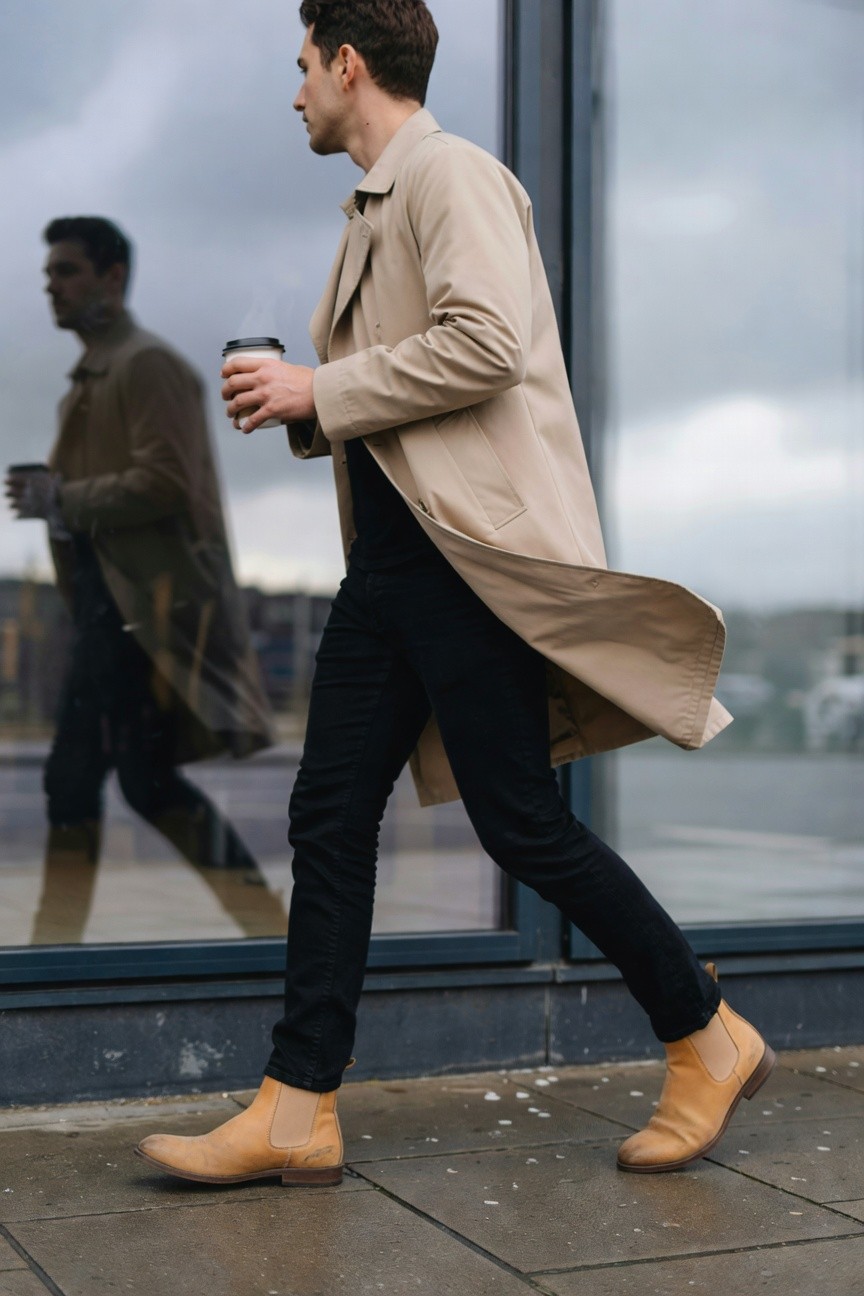 Side profile of a man walking in a long beige trench coat over slim black jeans and tan leather Chelsea boots, holding a coffee cup, reflected in a glass building