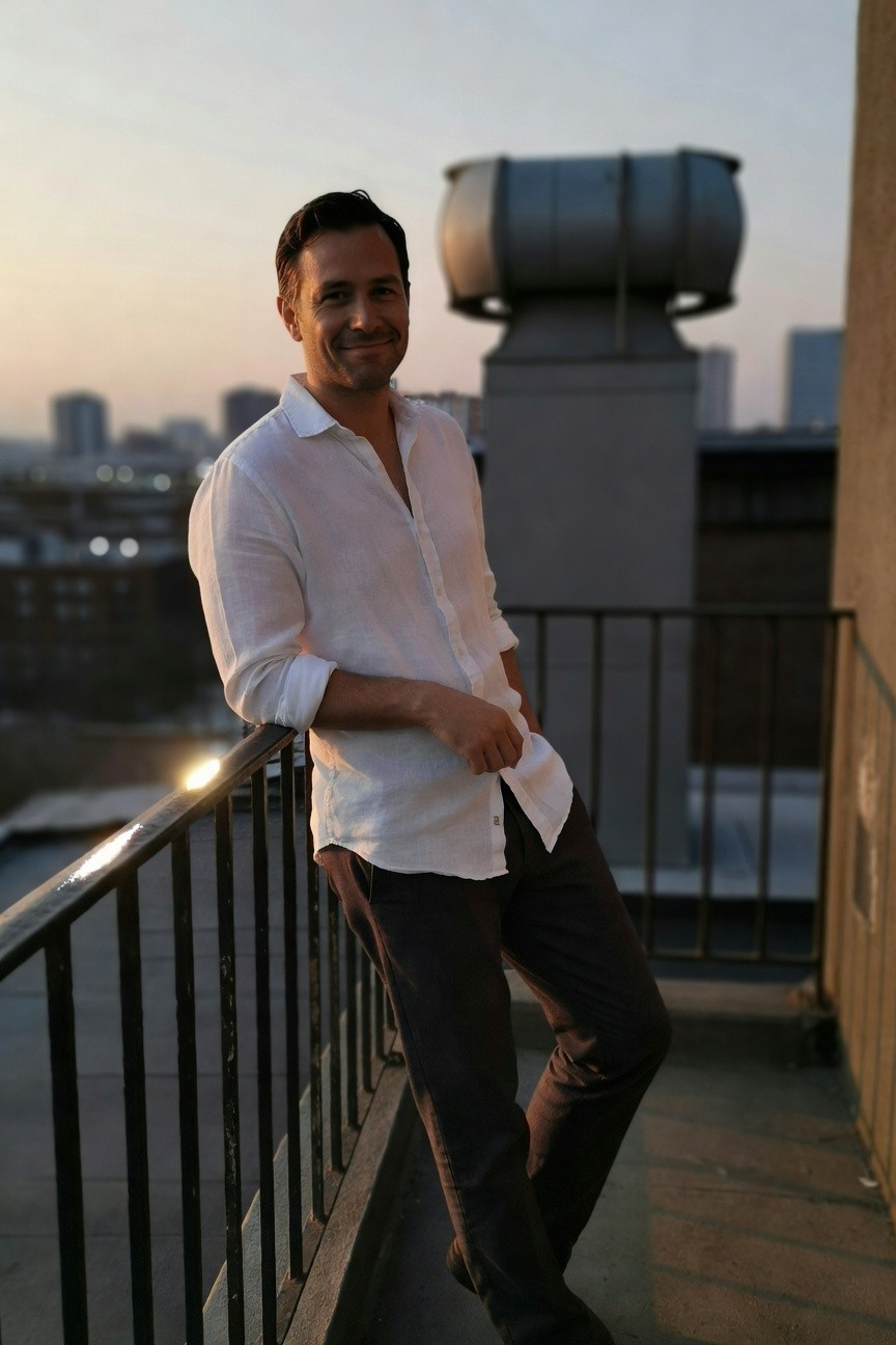 Man leaning on balcony railing in white linen shirt with open collar and grey dress pants, city buildings in background at dusk