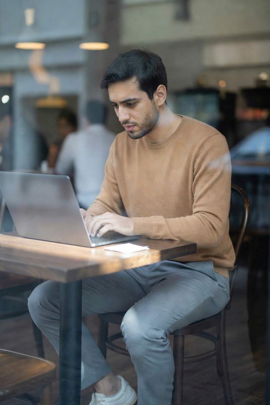 Man in beige knit sweater and light grey dress pants typing on laptop at wooden cafe table, white sneakers visible, focused office casual style