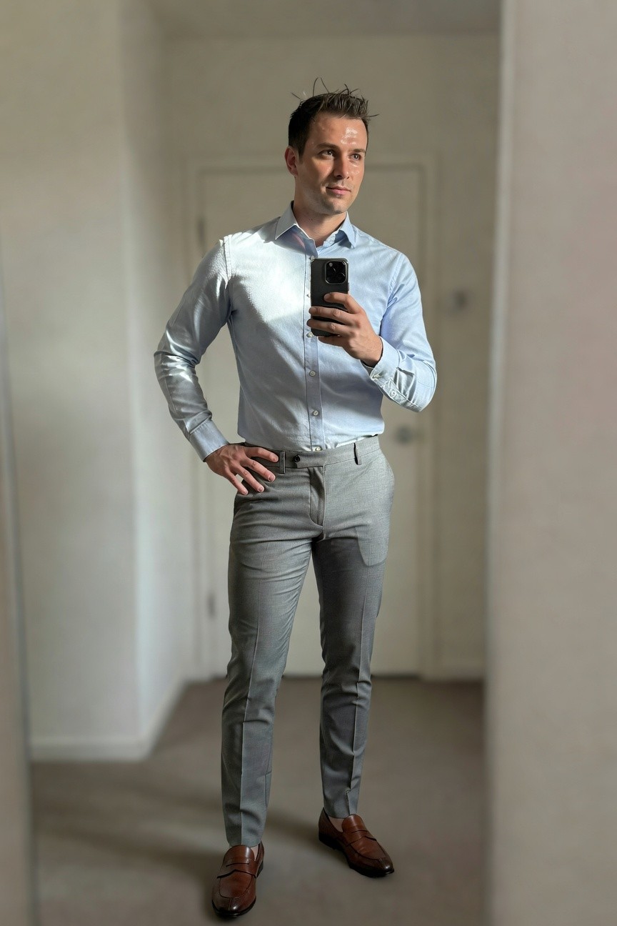 Man in light blue long-sleeve dress shirt tucked into slim grey dress pants with brown leather loafers, mirror selfie in hallway showing full professional office outfit