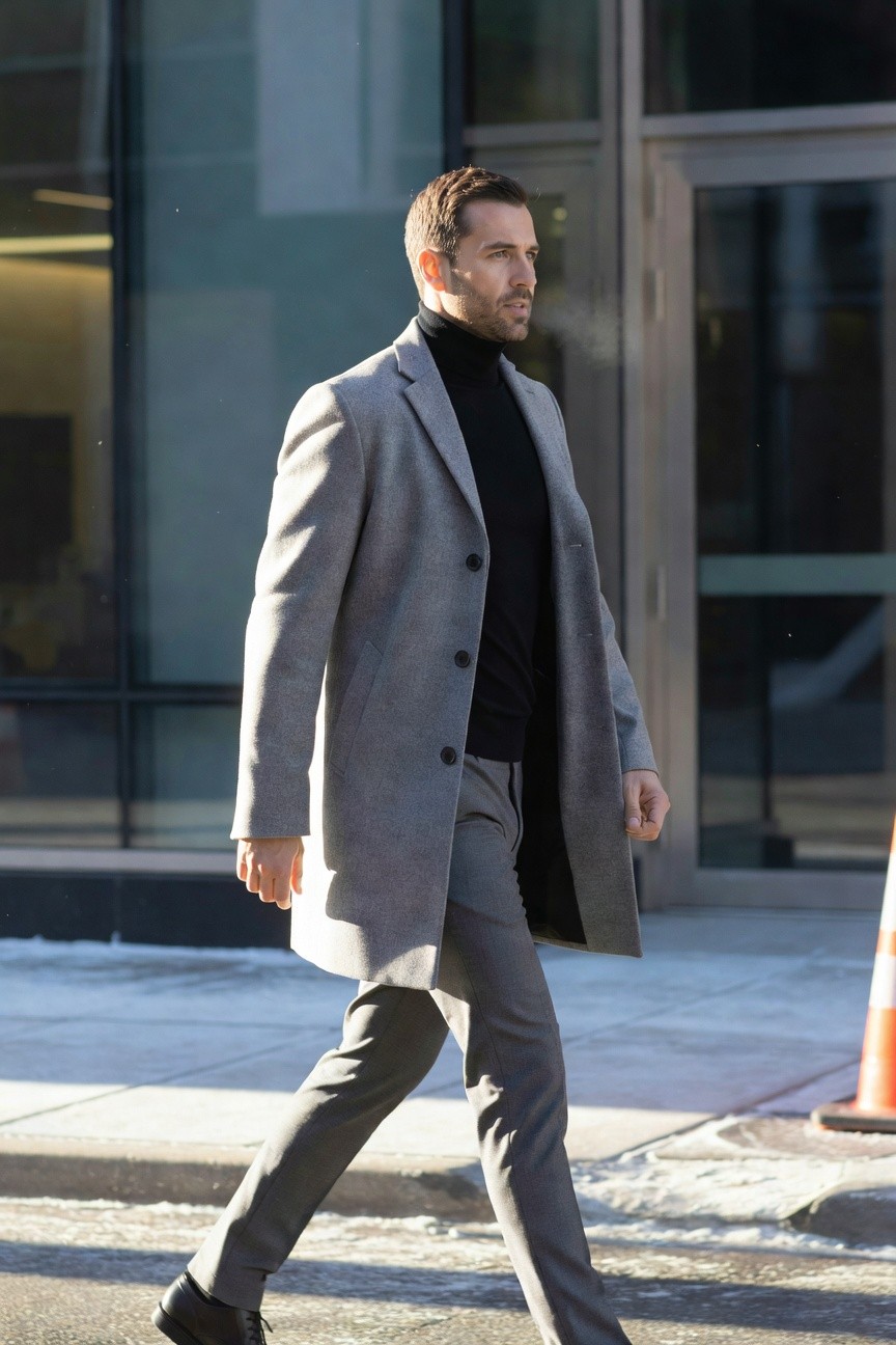 Man walking in tailored long grey wool overcoat layered over black turtleneck sweater with slim grey dress pants and black leather oxford shoes