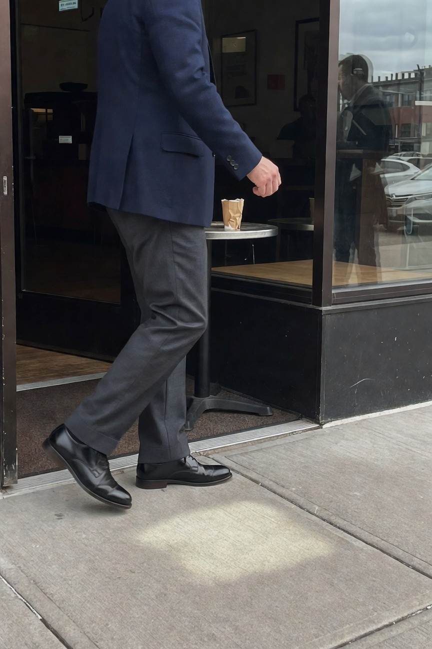 Side view of a man in navy blazer, grey dress pants, and black loafers stepping out a door, holding a coffee cup, sharp office outfit