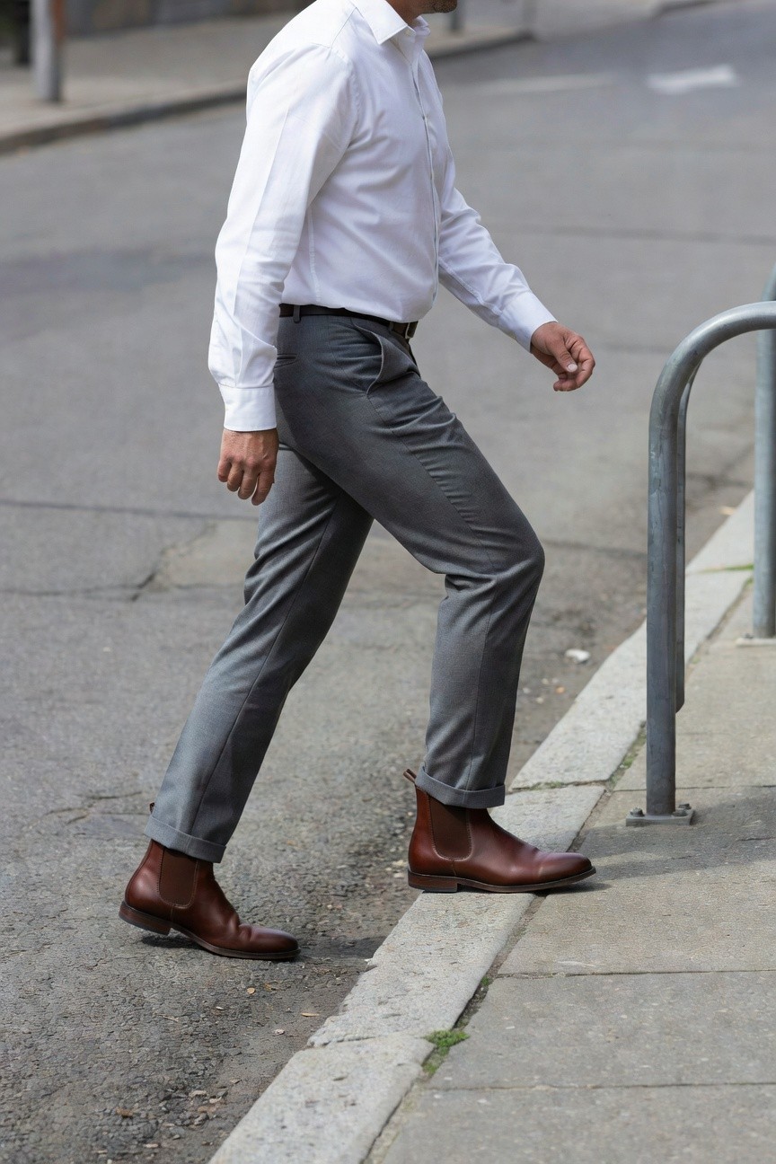 Side view of man in slim grey dress pants cuffed above brown leather Chelsea boots, white button-up shirt, walking on urban sidewalk near metal railing