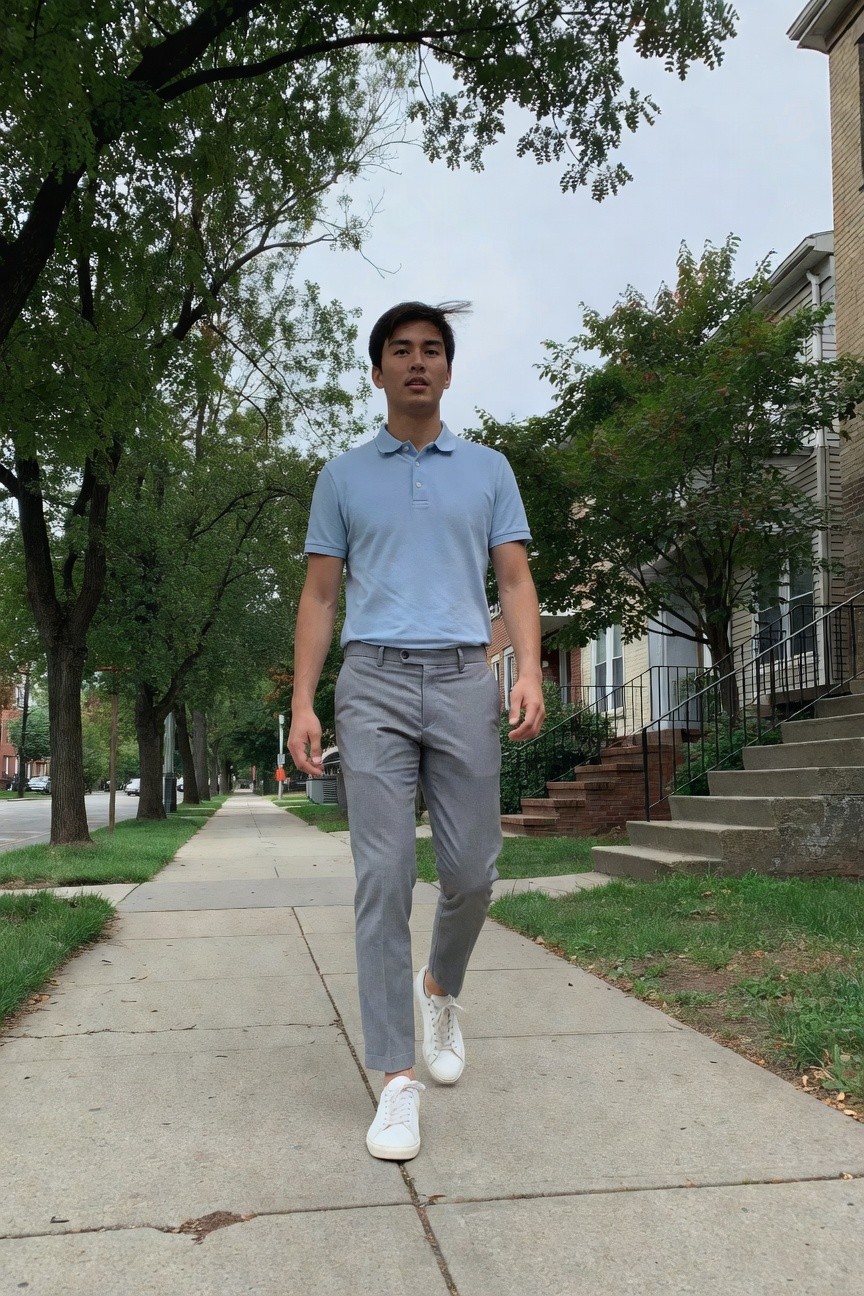 A man walking on a sidewalk wearing a light blue short-sleeve polo shirt, slim grey trousers, and white sneakers, captured mid-stride in a tree-lined urban street.