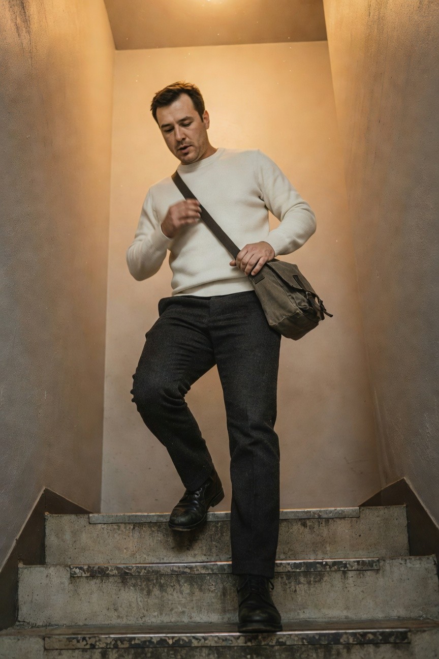 Man ascending concrete stairs in cream knit sweater, dark grey wool trousers, khaki canvas messenger bag with leather strap, and black leather boots, warm indoor lighting