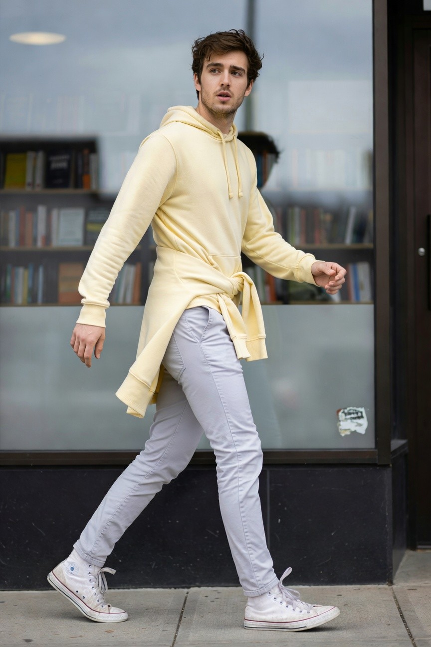 A man in a long pale yellow asymmetric hoodie with side tie, light grey slim pants, and white Converse high-tops, walking casually outside a bookstore window
