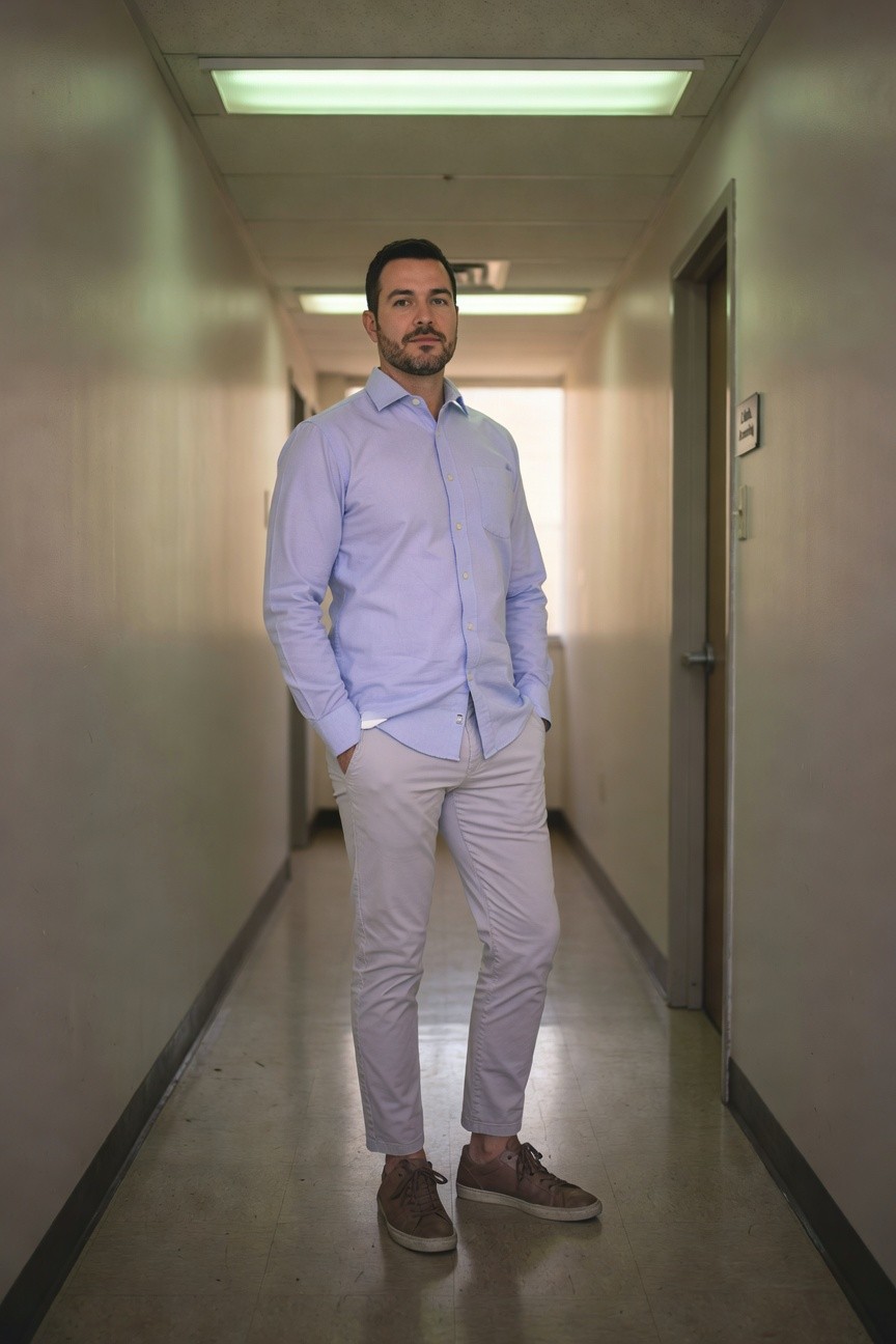 Man in a beige hallway wearing light blue chambray button-up shirt with sleeves rolled, light grey slim cropped pants, hands in pockets, brown suede sneakers, casual professional stance