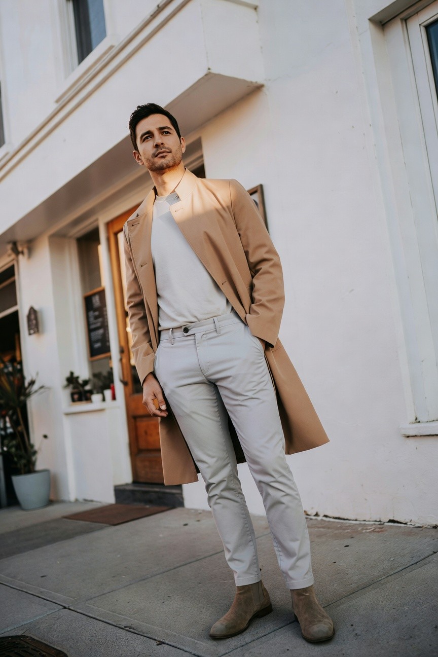 Man in light grey pleated pants, white t-shirt, open long camel overcoat, and beige suede chelsea boots, posing casually with hands in pockets