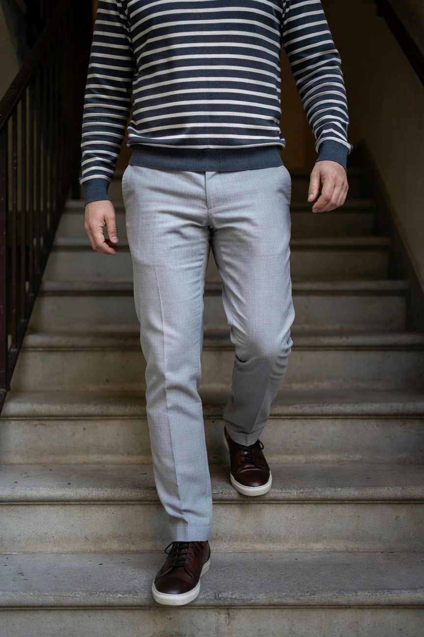 Man walking up stairs in navy and white horizontal striped knit sweater with grey ribbed neckline, light grey slim trousers, and brown leather lace-up sneakers