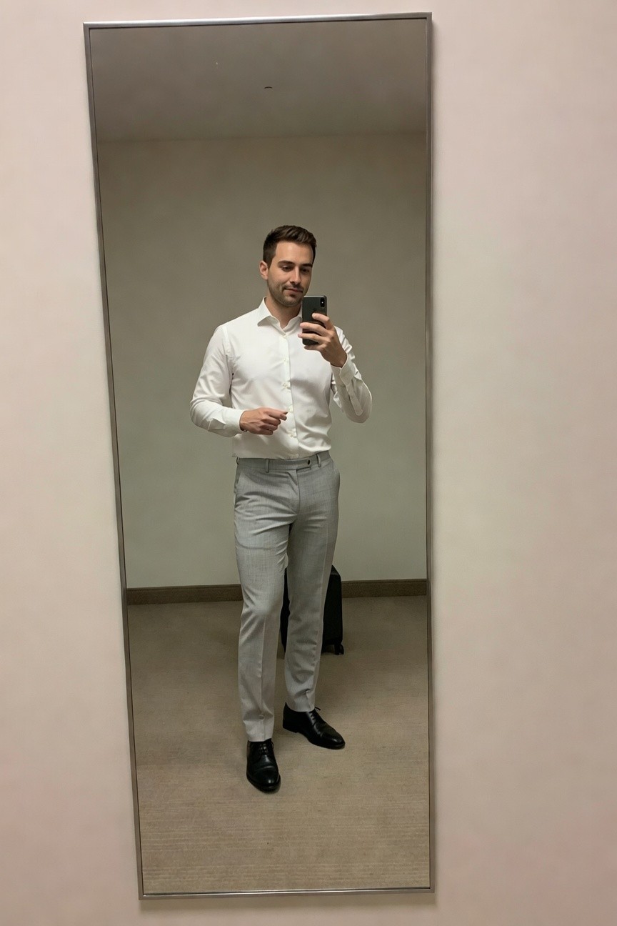 Man taking mirror selfie in white long-sleeve button-up shirt tucked into slim light grey trousers with black dress shoes and grey briefcase nearby in neutral room