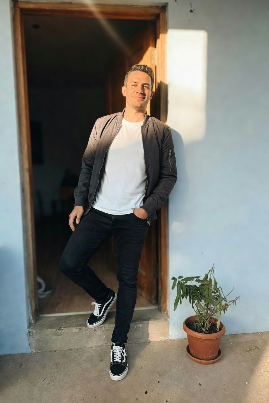 Man posing casually in open doorway wearing gradient gray bomber jacket over white t-shirt tucked into slim black jeans and black Vans sneakers with white soles, one foot propped up against doorframe next to potted plant