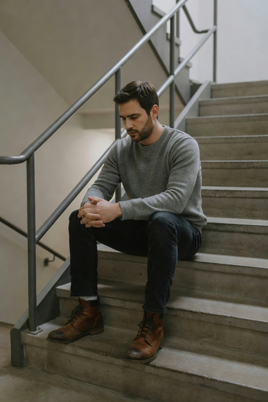 Man seated on concrete stairs in gray crewneck sweater, slim dark jeans rolled at ankles, brown laced leather boots, hands clasped pensively