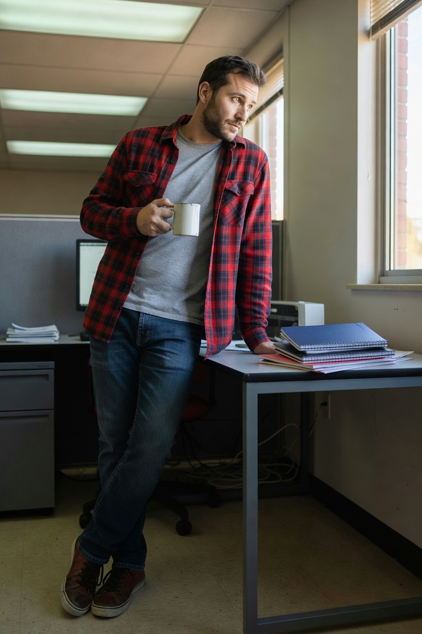 Man in open red-and-black plaid flannel shirt over gray t-shirt, blue straight-leg jeans, and brown high-top sneakers, leaning casually against office desk while holding white coffee mug