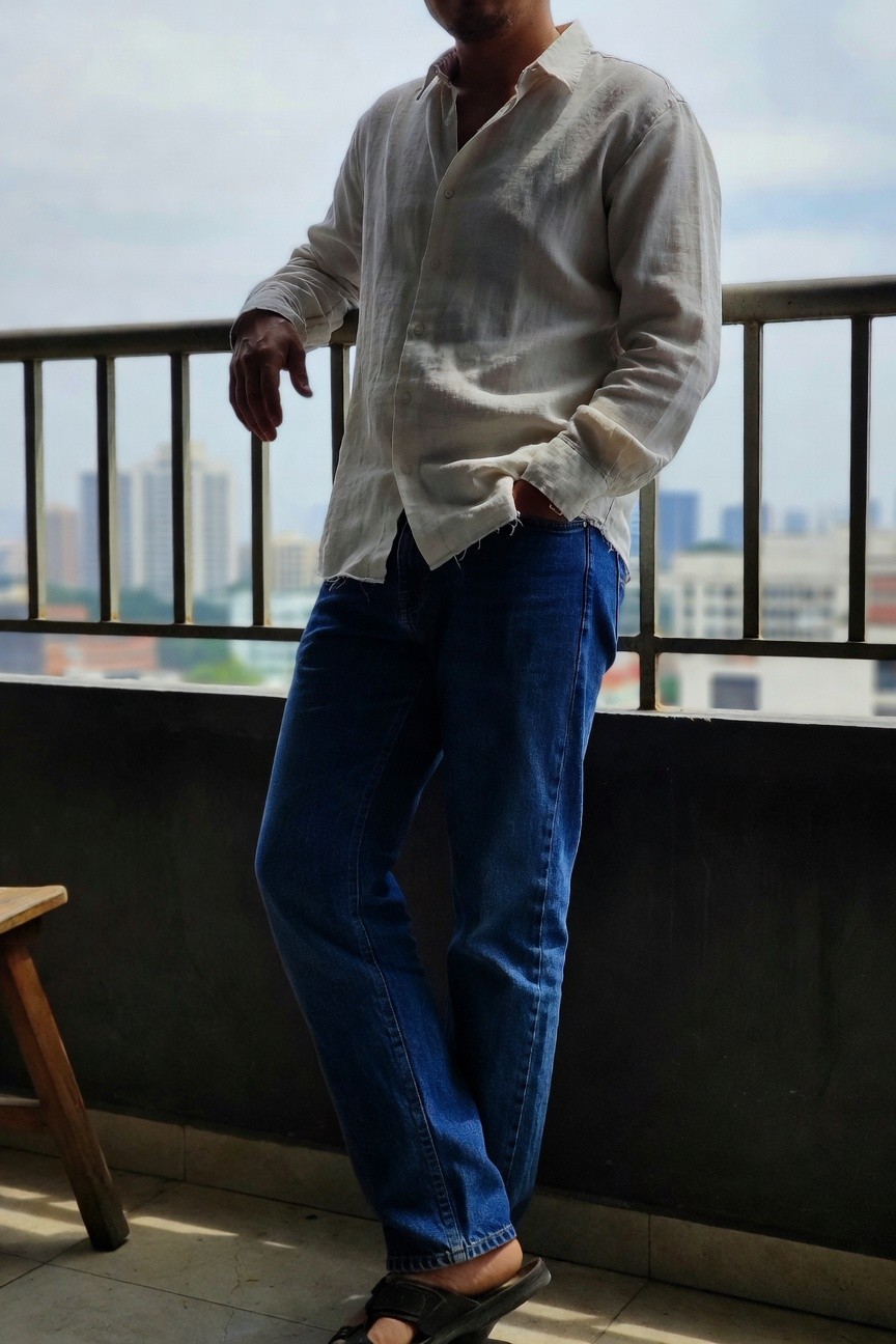 Man in a loose white linen shirt unbuttoned at the top over straight-leg blue jeans and black sandals, leaning casually against a balcony railing