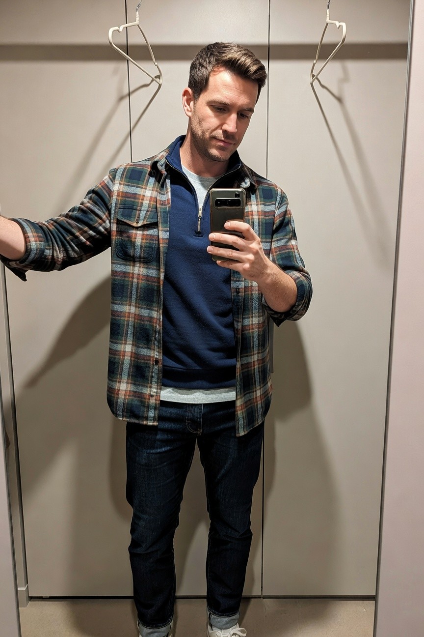 Man in a mirror selfie wearing an open navy-and-rust plaid flannel shirt over a navy quarter zip pullover, white crewneck tee, slim dark blue jeans cuffed at the ankles, and white sneakers with blue accents, arms extended while holding a phone.