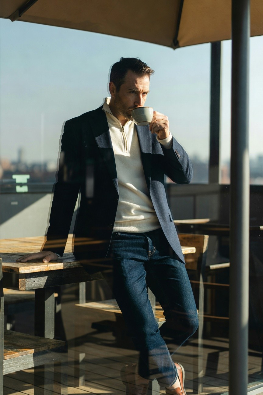 Man in navy pinstripe blazer over cream quarter zip sweater, dark slim jeans, and tan loafers, seated casually at a wooden table sipping from a white mug