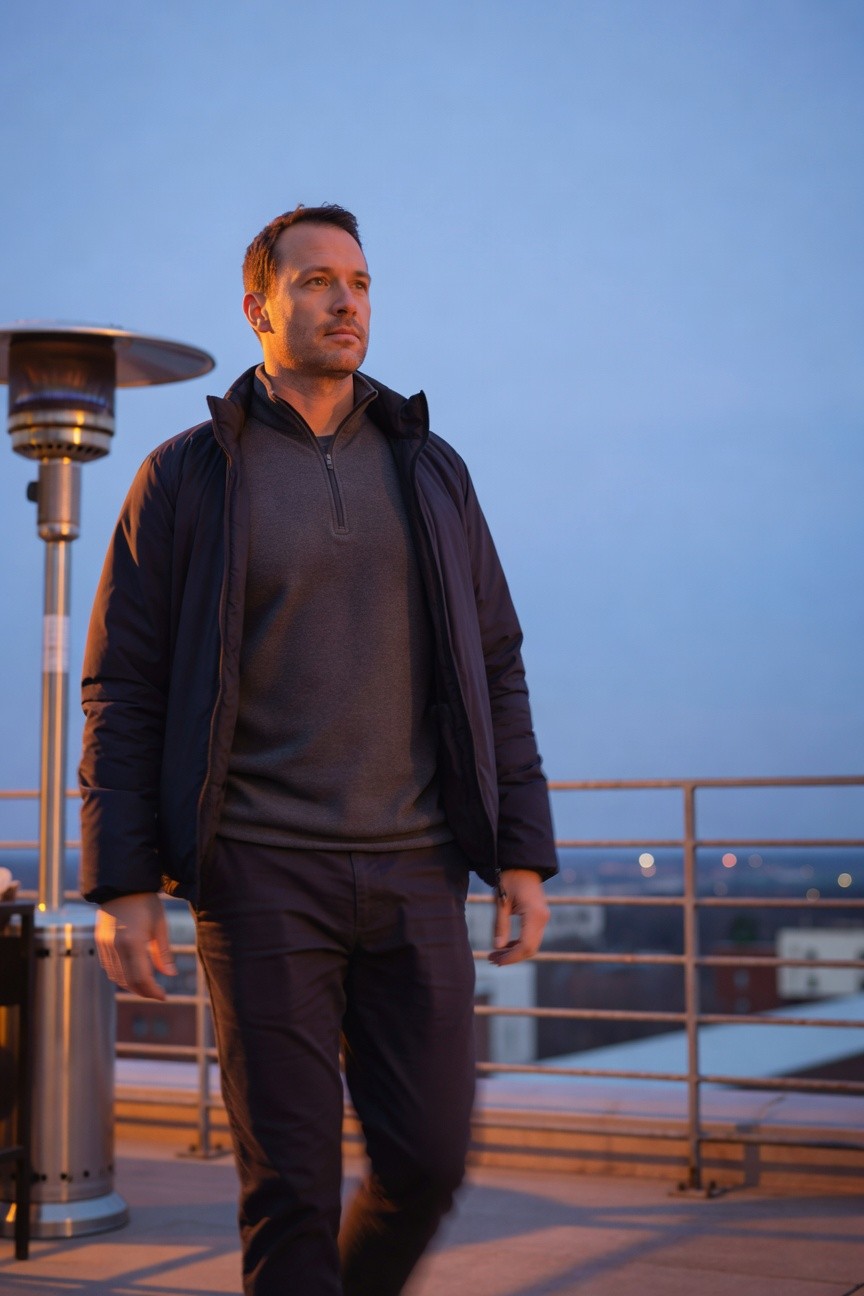 Man in dark charcoal quarter-zip sweater layered under open black zip-up jacket and slim dark trousers, standing on rooftop near patio heater at dusk