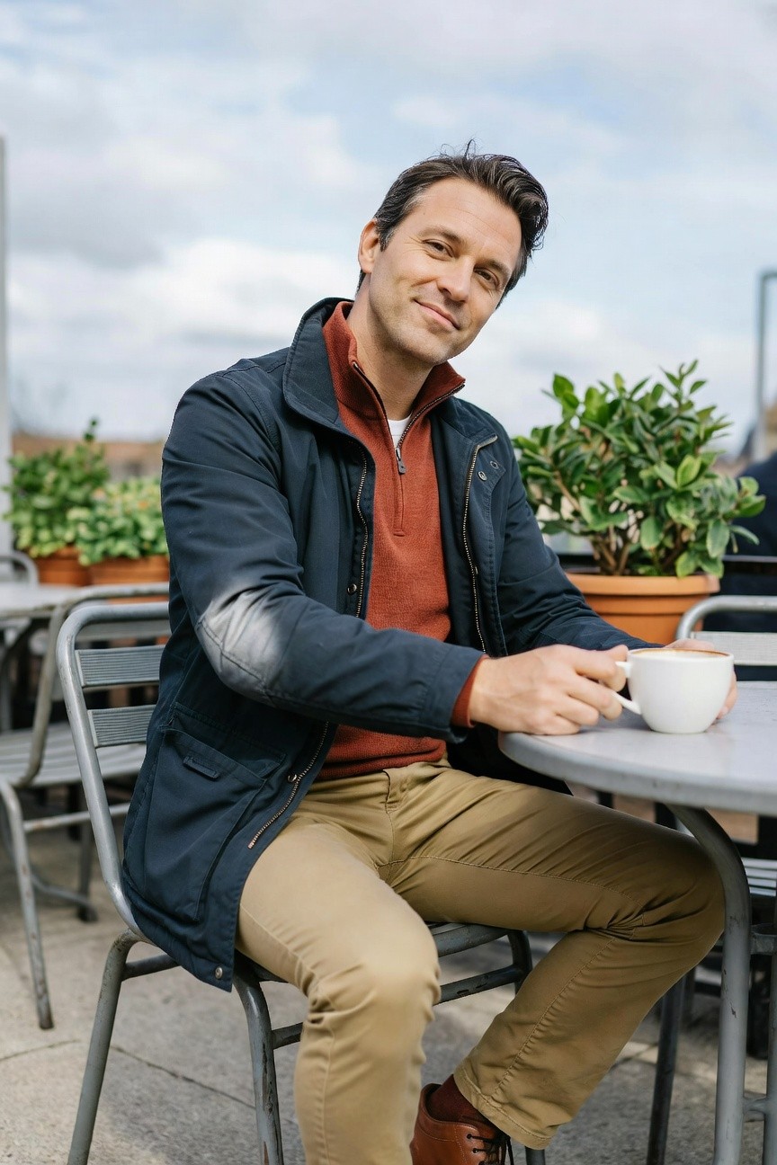 Man in rust quarter zip sweater layered under navy waxed field jacket with elbow patches, khaki chinos, and brown leather brogues, sitting at outdoor table holding coffee mug