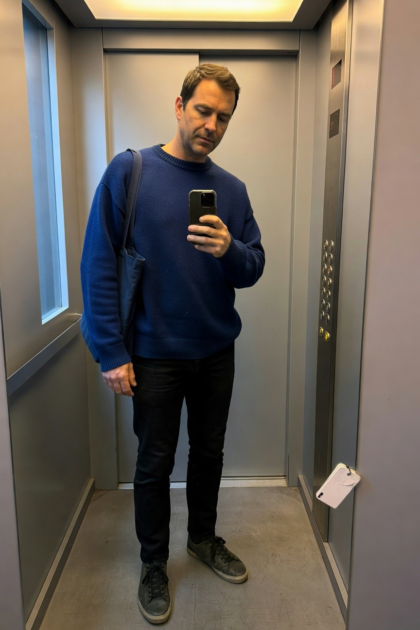 Man taking elevator selfie in oversized navy blue knit sweater, black shoulder bag, slim black jeans, and grey sneakers with black accents