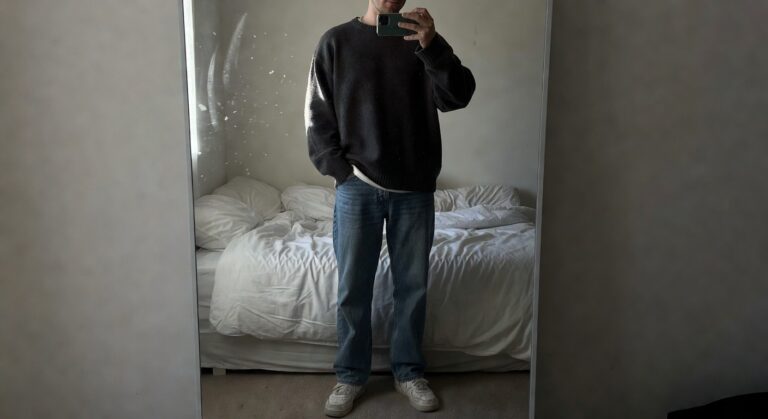 Man in oversized dark gray knit sweater, relaxed straight-leg blue jeans, and white low-top sneakers, taking a mirror selfie in a bedroom with hands in pockets