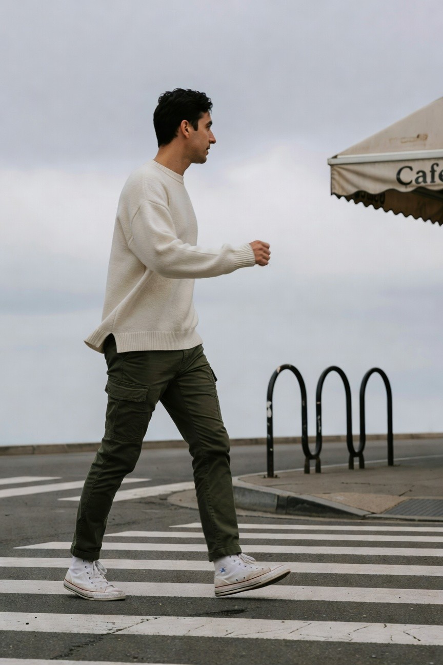 Side profile of a man walking in a cream oversized knit sweater, olive green cargo pants, and white Converse high-tops, captured mid-stride on a crosswalk near a cafe awning
