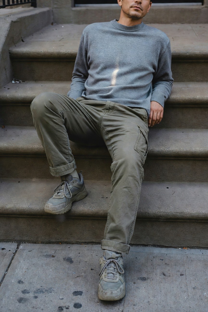 A man lounging on stone steps in a light gray oversized crewneck sweater, olive green cargo pants cuffed at the ankles, and worn gray chunky sneakers with white accents, arms relaxed at his sides