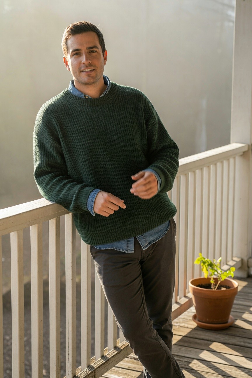 Man in oversized green ribbed knit sweater layered over a blue button-down shirt with grey trousers, leaning casually on a wooden railing with hands in relaxed pose