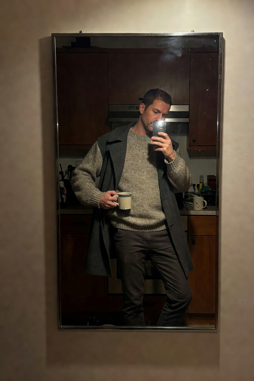 Mirror selfie of man in chunky gray oversized knit sweater layered under dark gray coat, slim dark pants, wristwatch, holding white mug with blue rim, kitchen cabinets in background