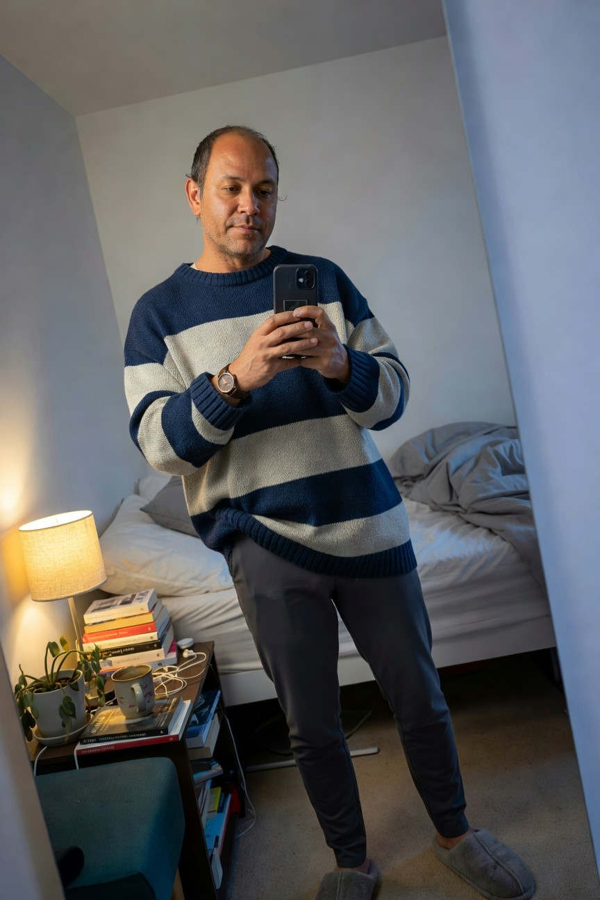 Man taking a mirror selfie in a bedroom, wearing a navy and white horizontal striped oversized knit sweater, slim grey jogger pants, grey slippers, brown leather watch, holding an iPhone, bedside table with lamp, books, plant, and mug nearby