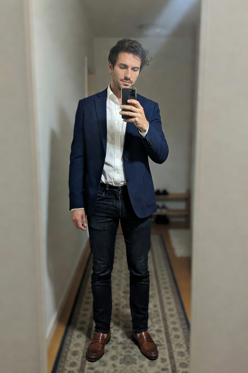 Mirror selfie of man in navy blue single-breasted blazer, white button-up shirt, slim dark wash jeans, and brown leather penny loafers, standing in hallway with shoe rack nearby