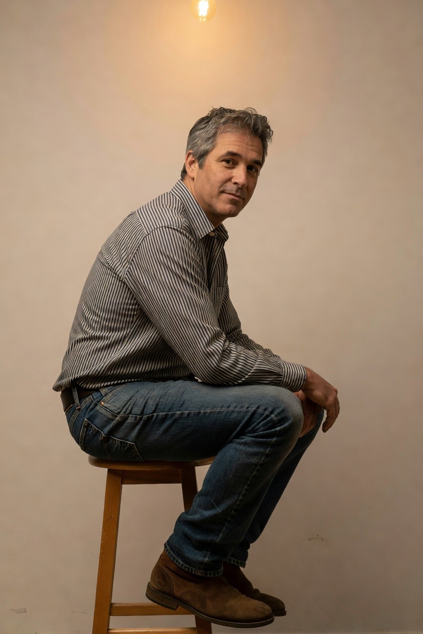 Side profile of a man in his 40s sitting on a wooden stool, wearing a gray and white striped long-sleeve button-up shirt tucked into dark blue straight-leg jeans, with brown leather ankle boots, arms crossed casually