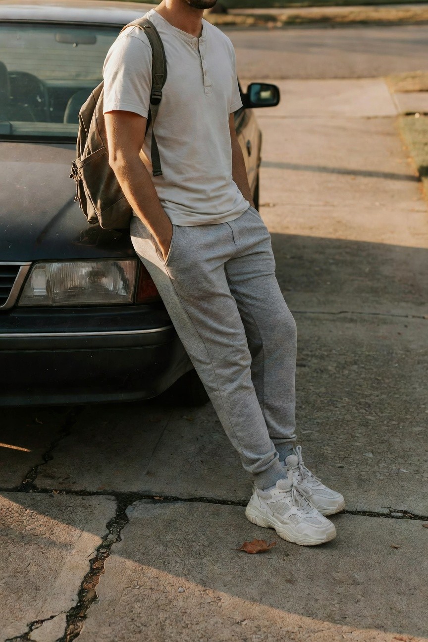 Man in cream short-sleeve henley shirt, grey jogger pants, white chunky platform sneakers, and olive green backpack, leaning casually with hands in pockets