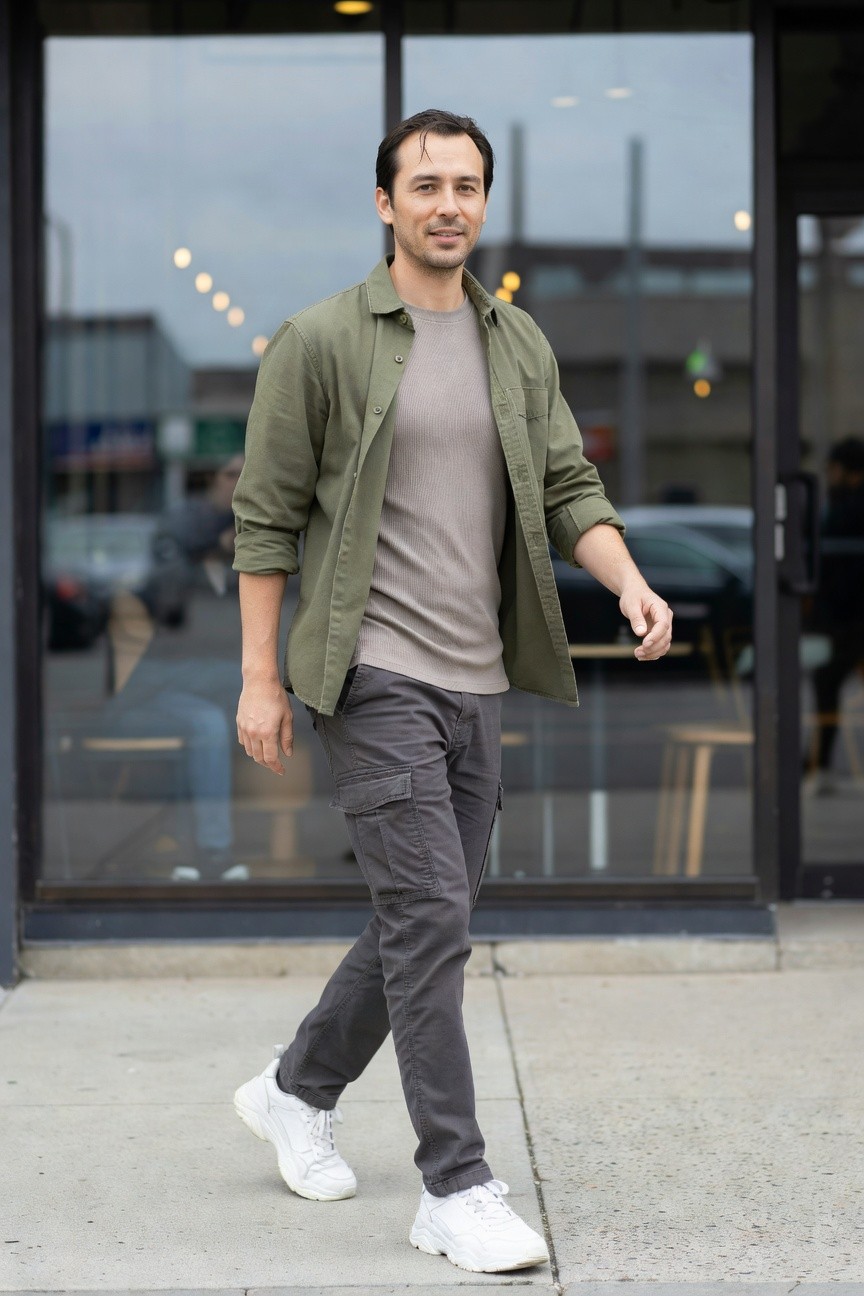 Man in open olive green cotton shirt jacket over grey t-shirt, charcoal grey cargo pants, and white chunky sneakers, walking casually toward glass doors