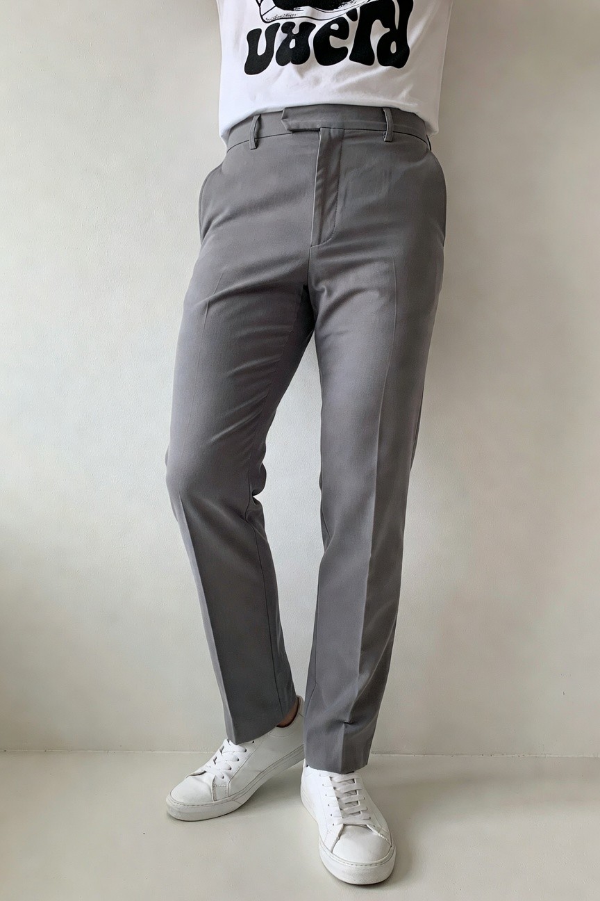 A man models slim light grey trousers with a slim fit, white cotton t-shirt featuring bold black 'UGLY NERD' graphic lettering, and white low-top sneakers, posed standing against a plain wall for a casual modern look