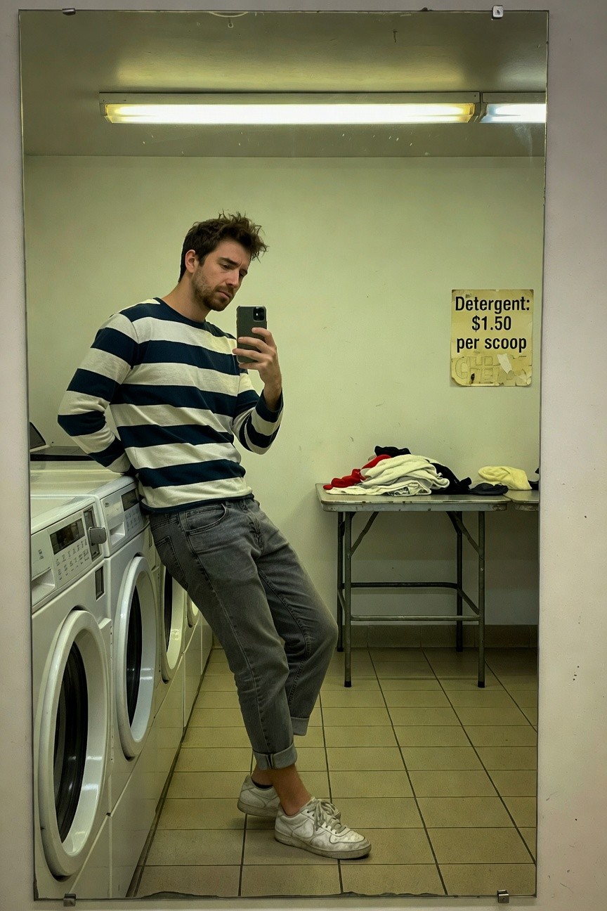 Man taking a mirror selfie in a laundromat, wearing a navy and white striped long-sleeve sweater, slim grey jeans rolled at the ankles, white sneakers with visible socks, leaning against washing machines.