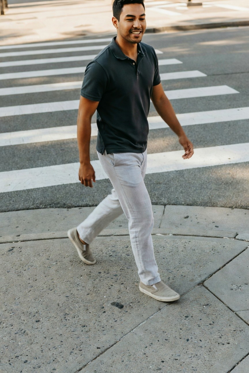 Man in dark navy short-sleeve polo shirt, light grey linen pants, and grey slip-on sneakers walking across a crosswalk, smiling casually
