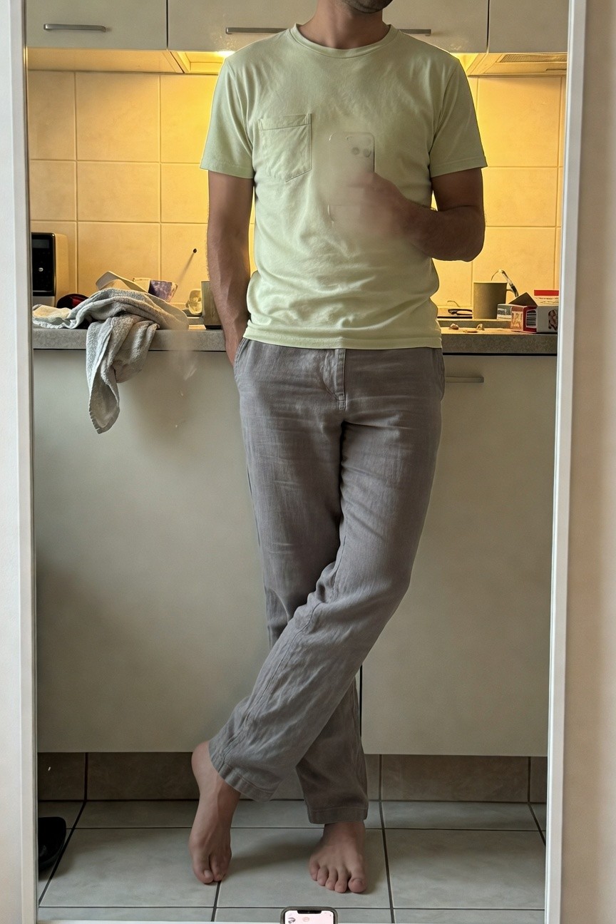 Man standing barefoot in a modern kitchen mirror selfie, wearing a light green short-sleeve pocket t-shirt and loose grey linen pants with hands in pockets, casual pose