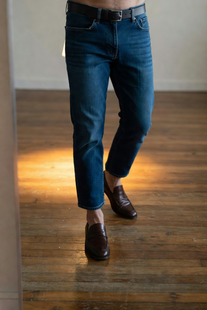 A man stands in slim dark blue ankle-length jeans with a black belt low on his hips, paired with brown leather loafers sockless, minimalist shirtless upper body for a clean sharp everyday outfit.