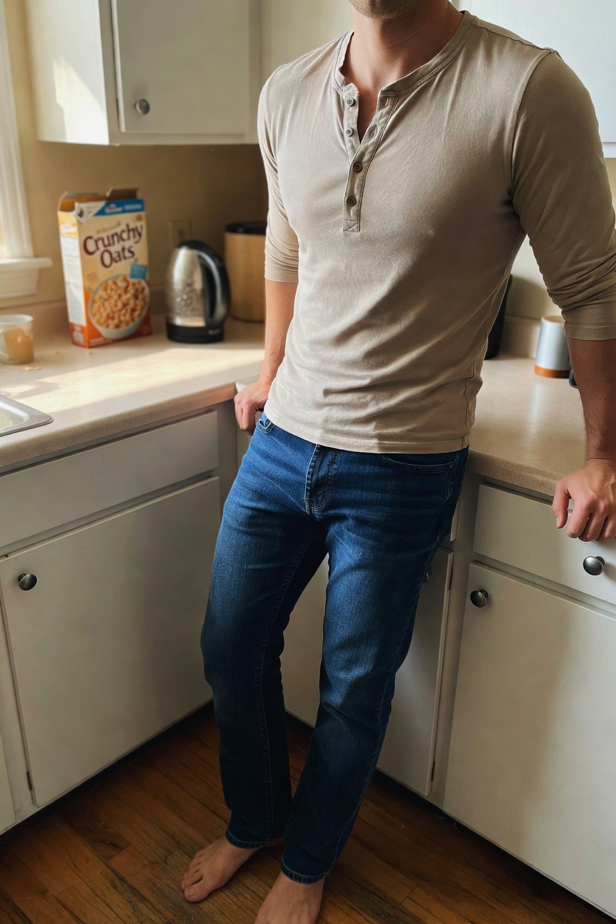 A man stands barefoot in a kitchen wearing a beige long-sleeve henley shirt with an open collar and dark blue straight-leg jeans, leaning casually against a white countertop with hands in pockets.
