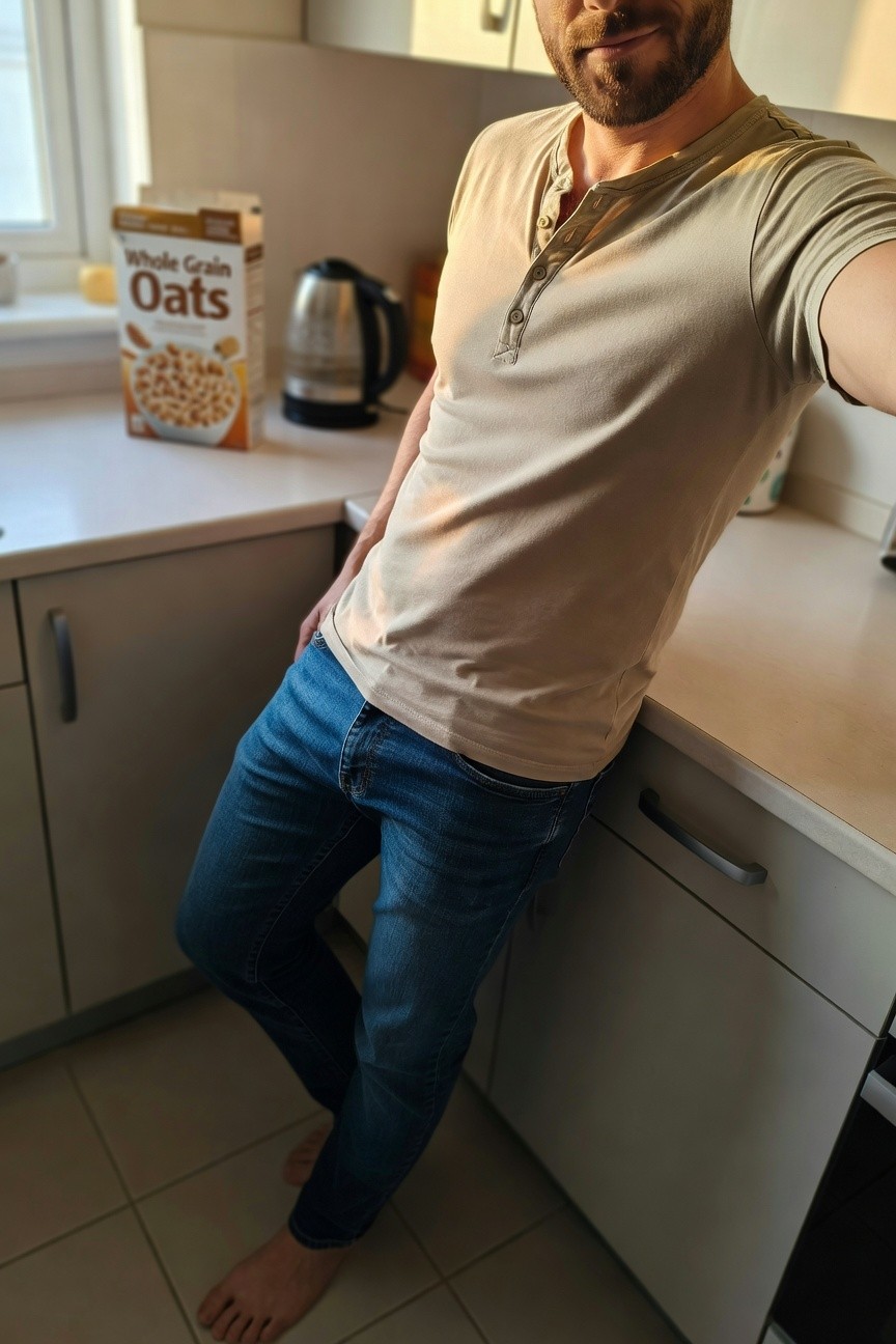 Bearded man in beige short-sleeve henley shirt and slim blue jeans leaning against white kitchen counter, barefoot on tile floor with box of oats nearby