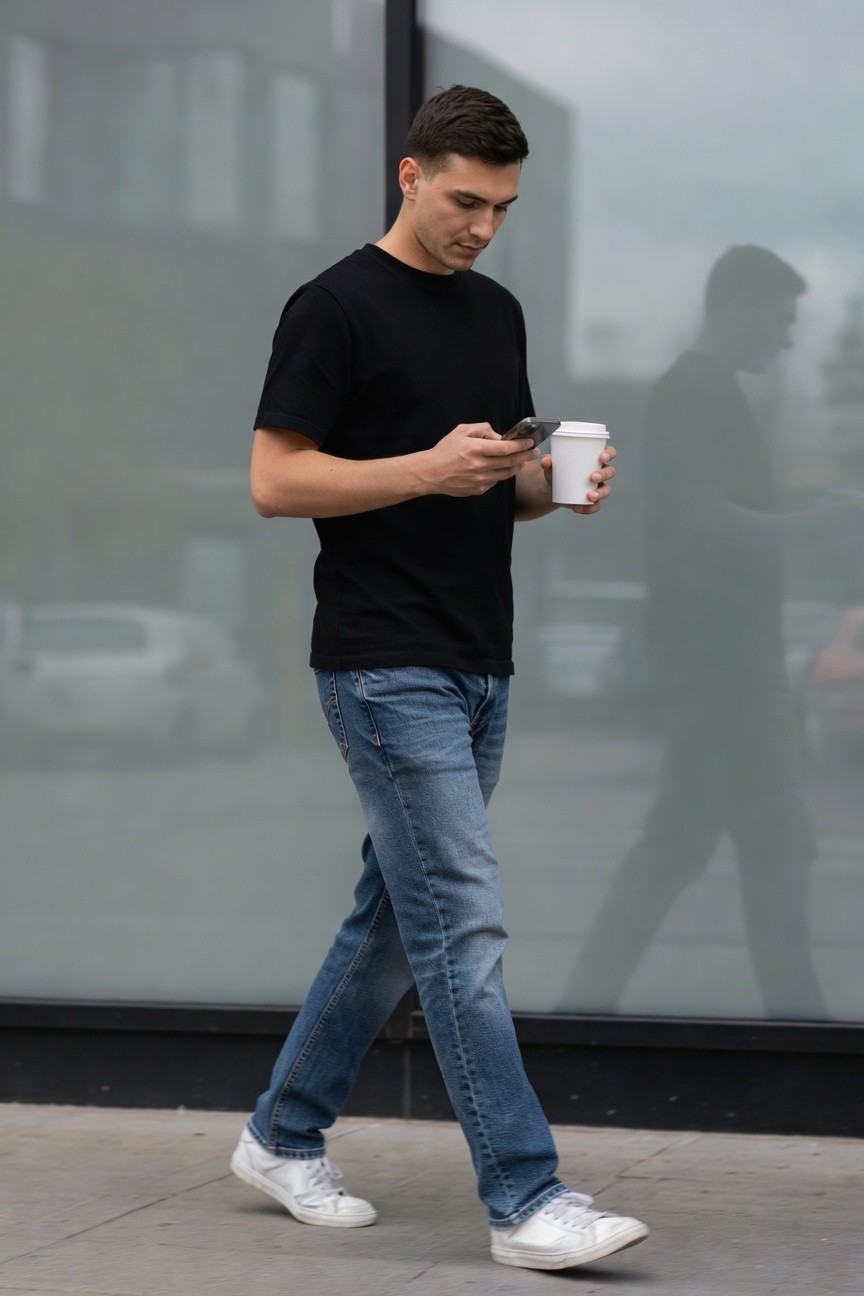 Side view of a man walking in a black short-sleeve t-shirt, straight blue jeans, and white sneakers, holding a smartphone in one hand and a white coffee cup in the other against a reflective glass building